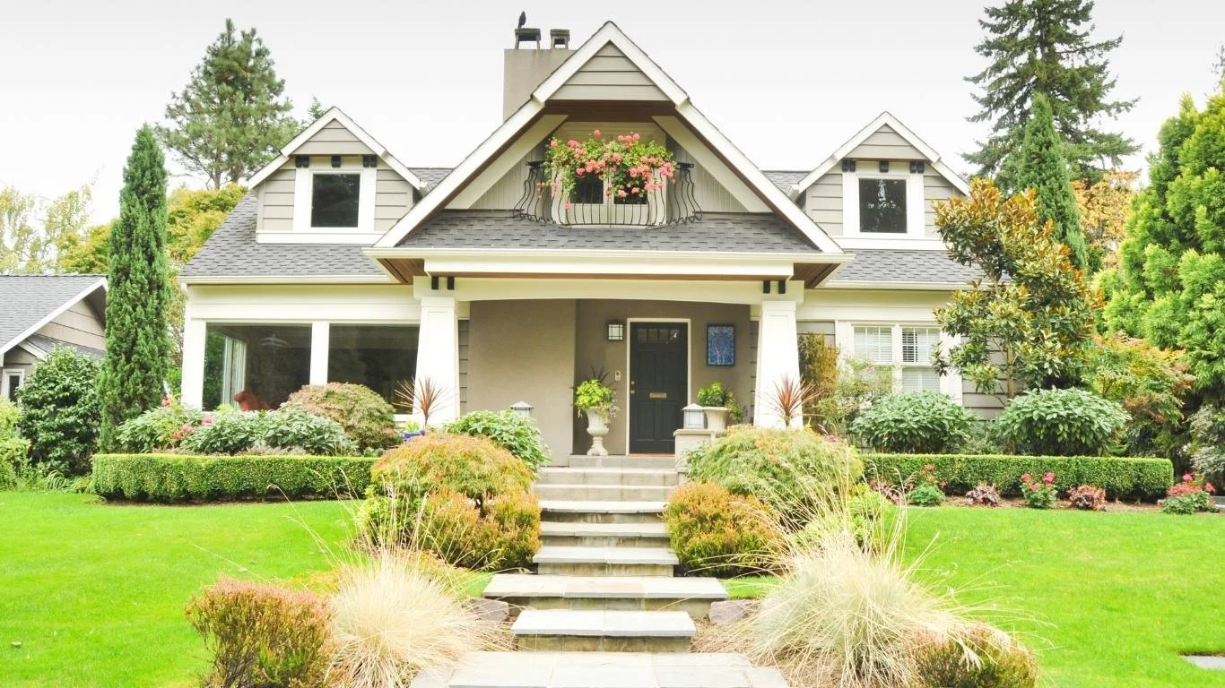 Front view of a gray two-story house with a gable roof, front steps, and landscaped front yard with bushes, flowers, and tall trees.