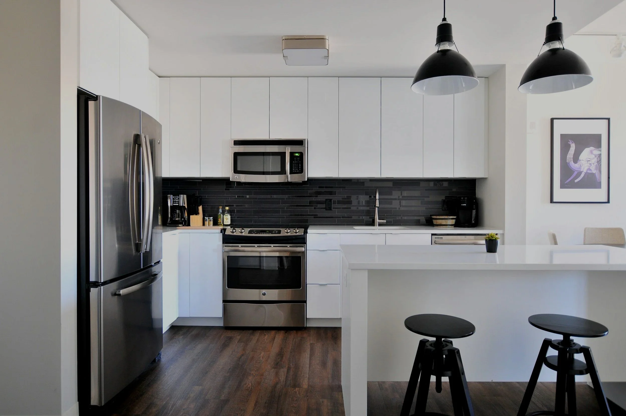 Modern kitchen with white cabinets, black backsplash, stainless steel refrigerator, microwave, stove, and sink. Two black pendant lights hang over the white breakfast bar with two black stools. Decor includes a small potted plant and framed artwork on the wall.