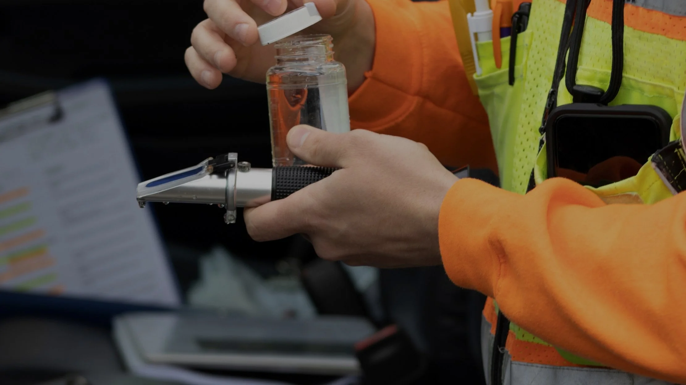 A person in orange and yellow safety clothing filling a small container with some clear liquid from a larger bottle, with a clipboard and other tools nearby.