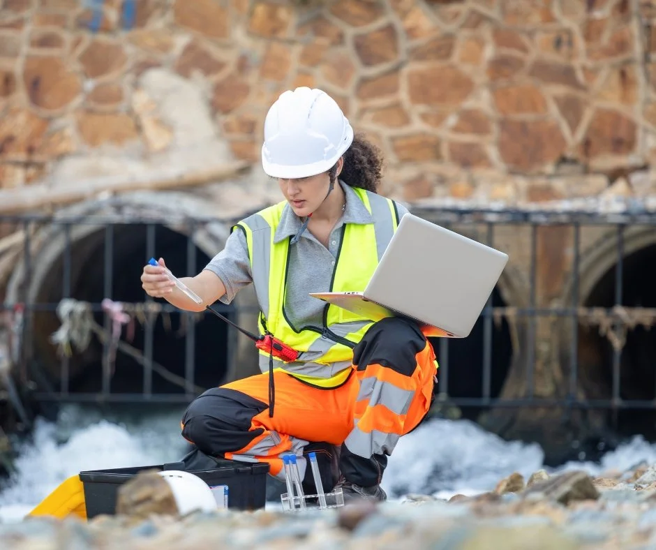 A female engineer wearing a white safety helmet, yellow safety vest, and orange safety pants, holding a laptop and testing equipment at a construction site with a stone wall and water pipes in the background.