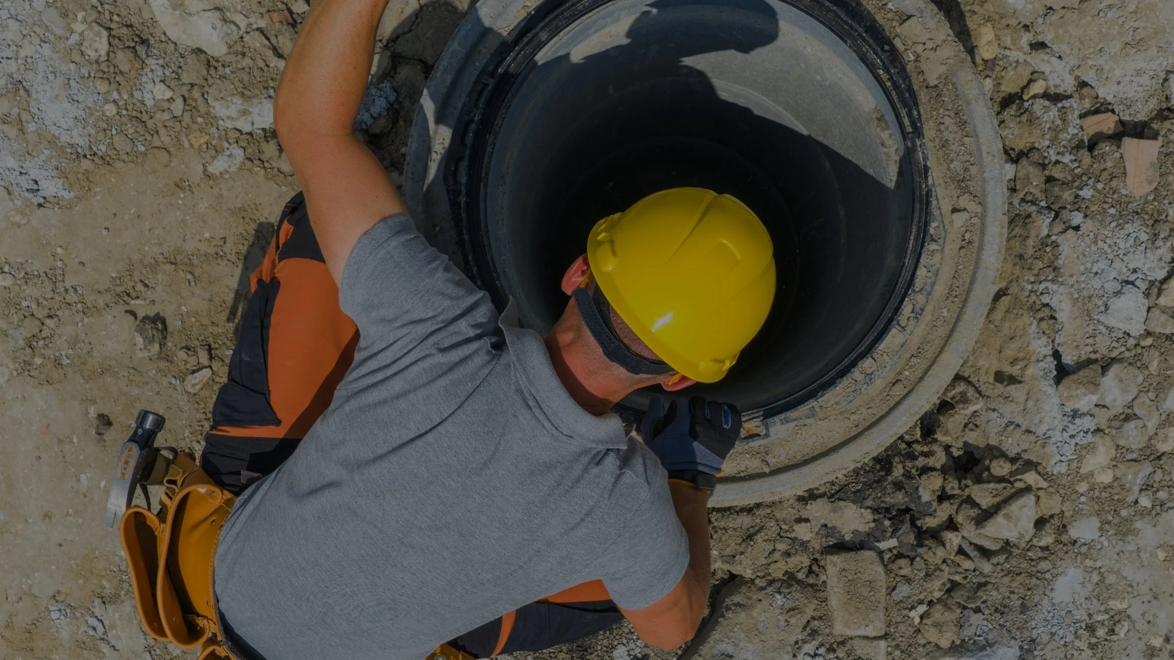 A construction worker wearing a yellow hard hat, gray shirt, and gloves working on a pipe in an open excavation site surrounded by dirt and rocks.