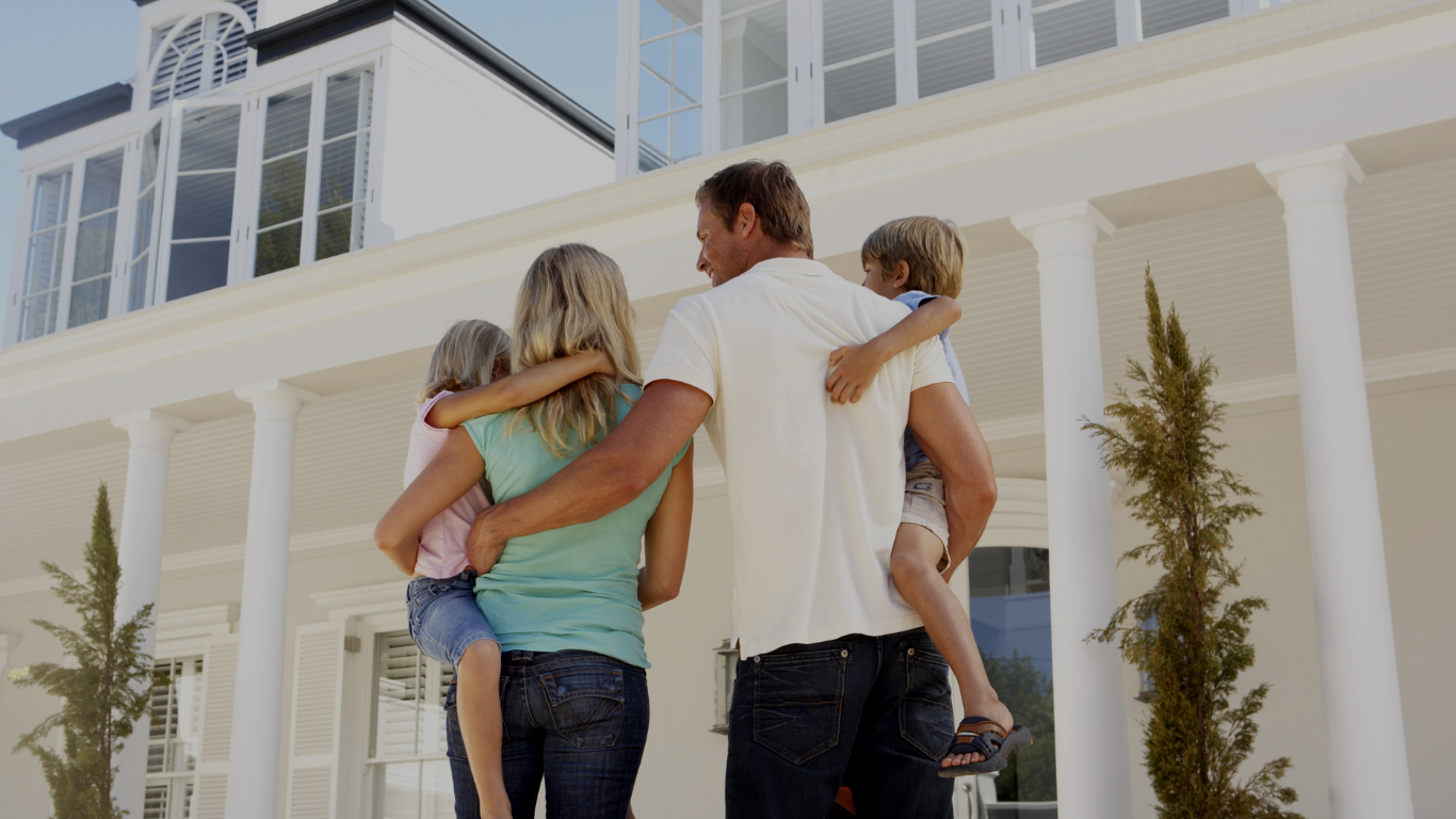 A family of four standing outside a large white house with columns. The parents are holding their children, who are hugging their parents.