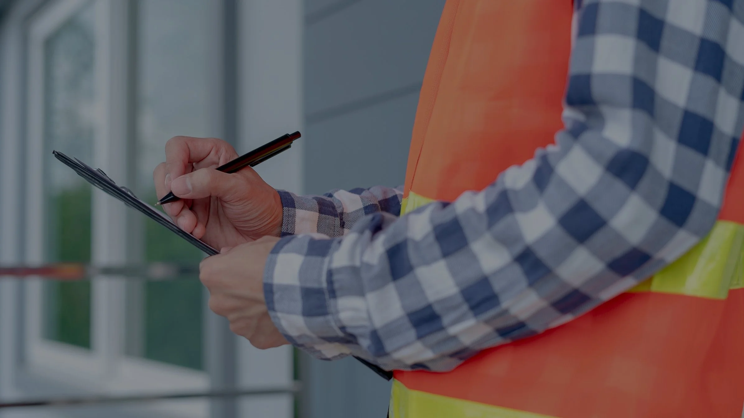 A construction worker in a plaid shirt and safety vest writing on a clipboard with a black pen inside a building or construction site.