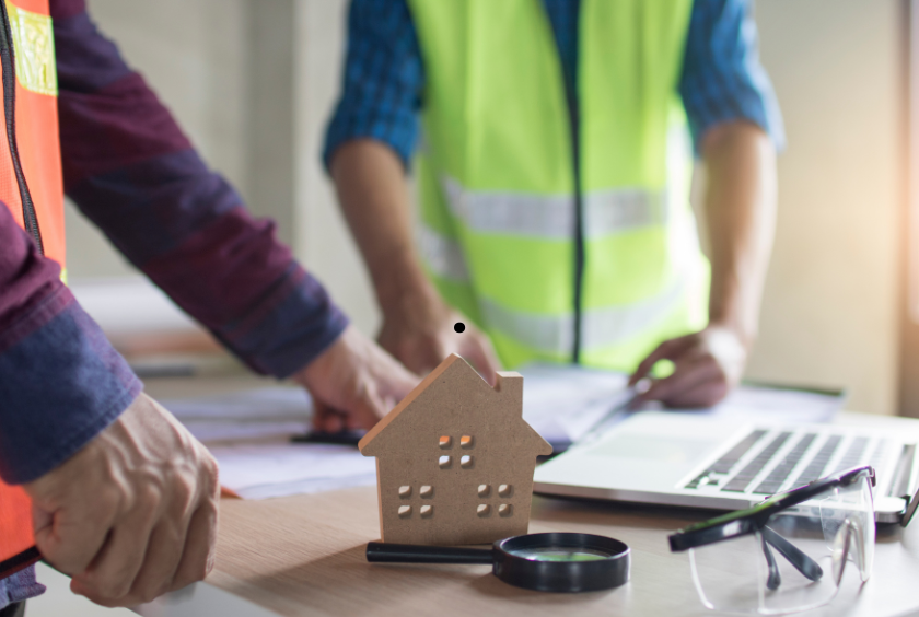 Two people in construction attire reviewing documents at a desk with a small house model, magnifying glass, and safety glasses.
