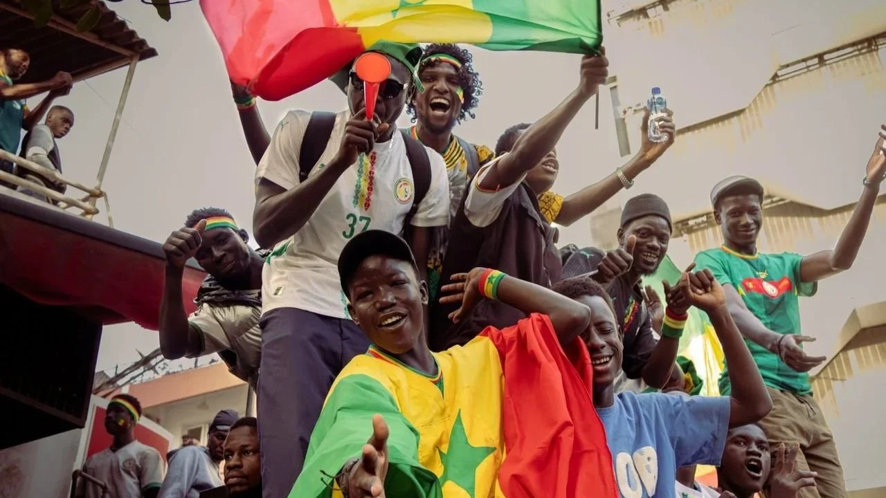 A joyful crowd of Senegalese sports fans cheering, smiling, and waving the green, yellow, and red flag of Senegal.
