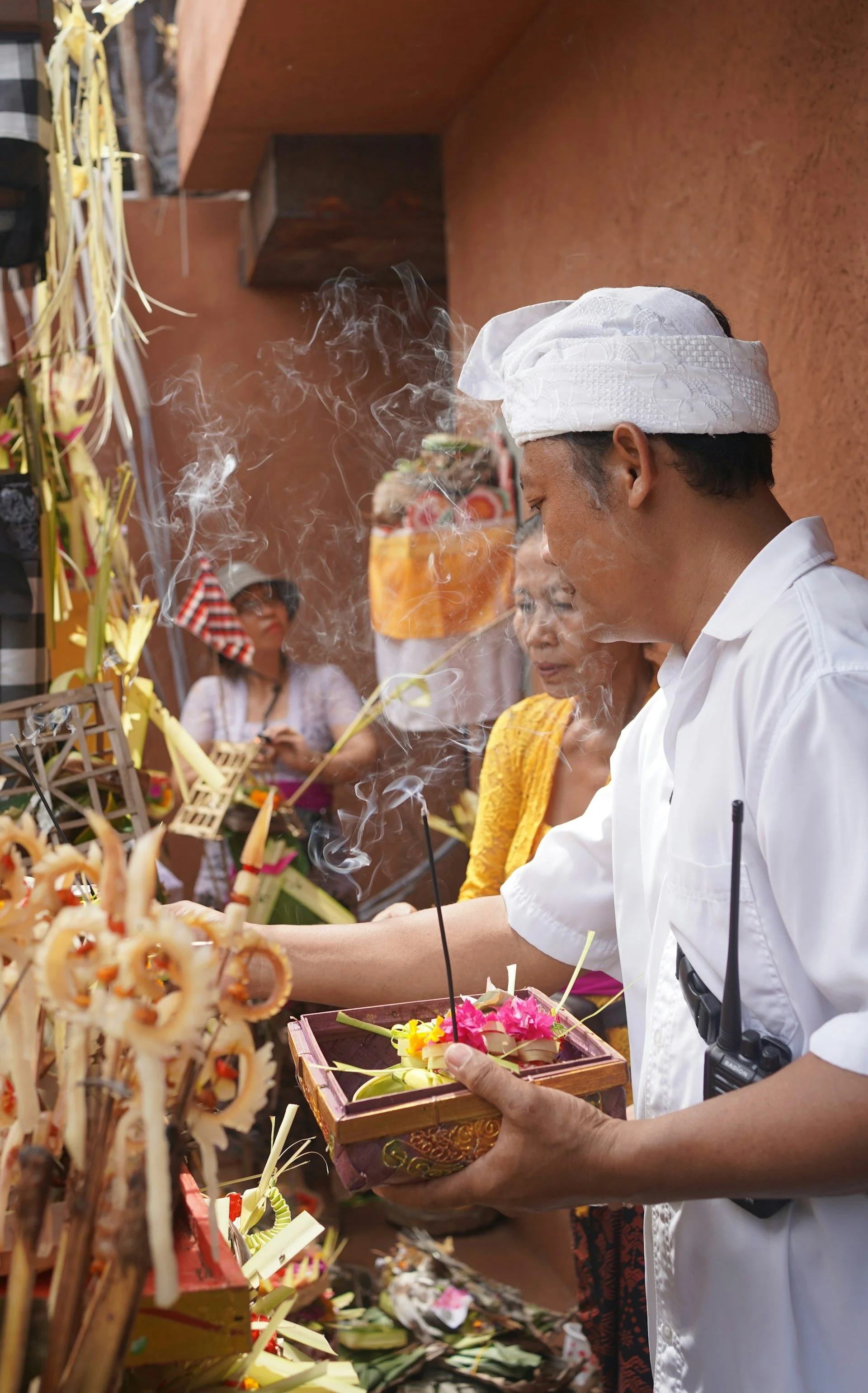 A Balinese man in traditional white ceremonial dress and headgear holding a tray of floral offerings amidst swirling incense smoke.