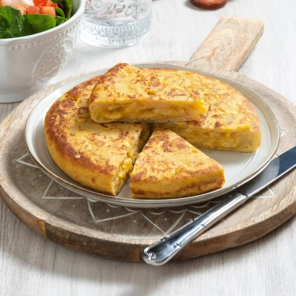 A sliced Spanish potato omelet (Tortilla Española) on a wooden board, served with a side salad, water, and chorizo sausage.