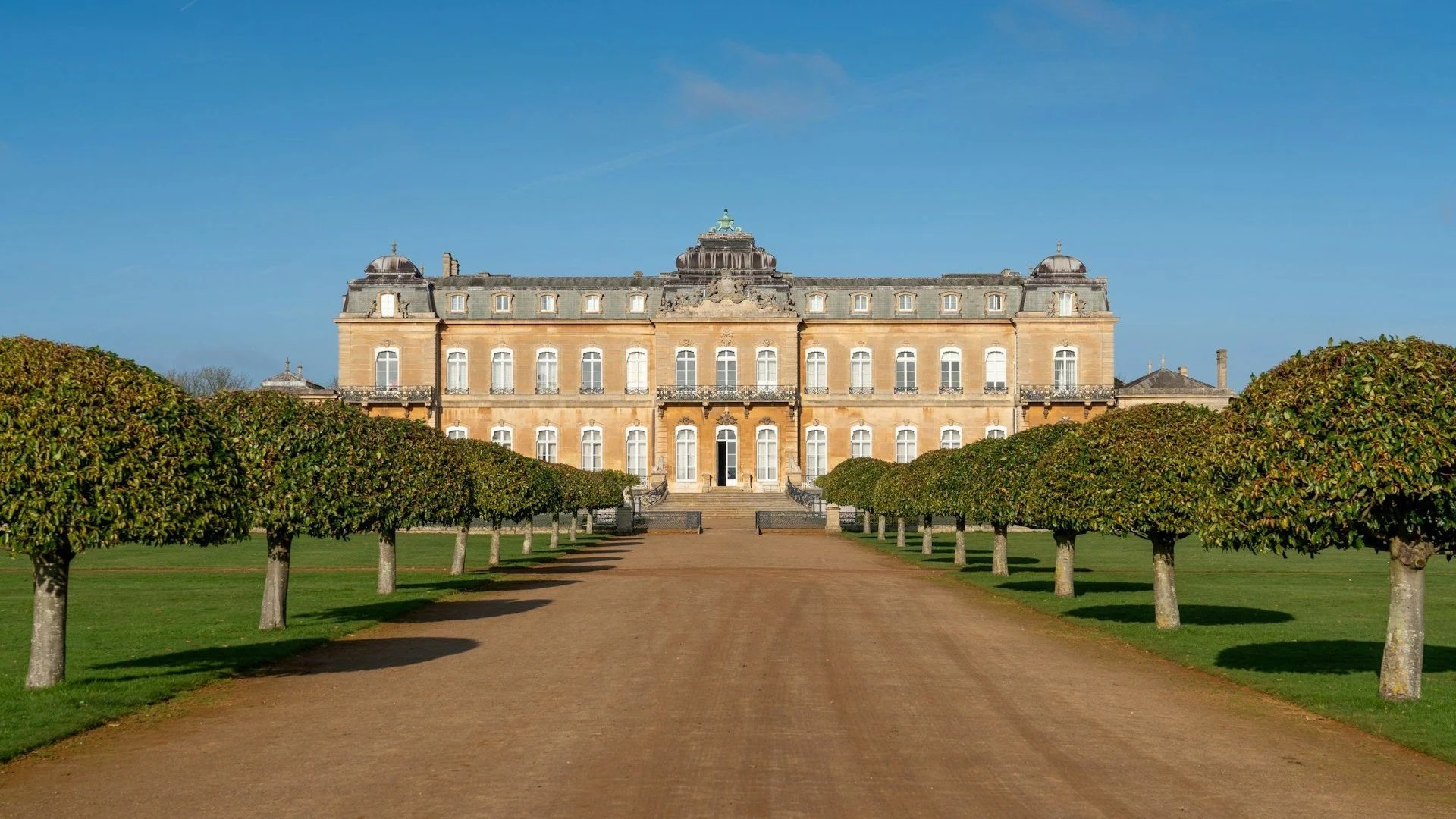 Gemini said A wide gravel path leads toward a grand, symmetrical honey-colored limestone mansion with ornate windows and a dark domed roof.