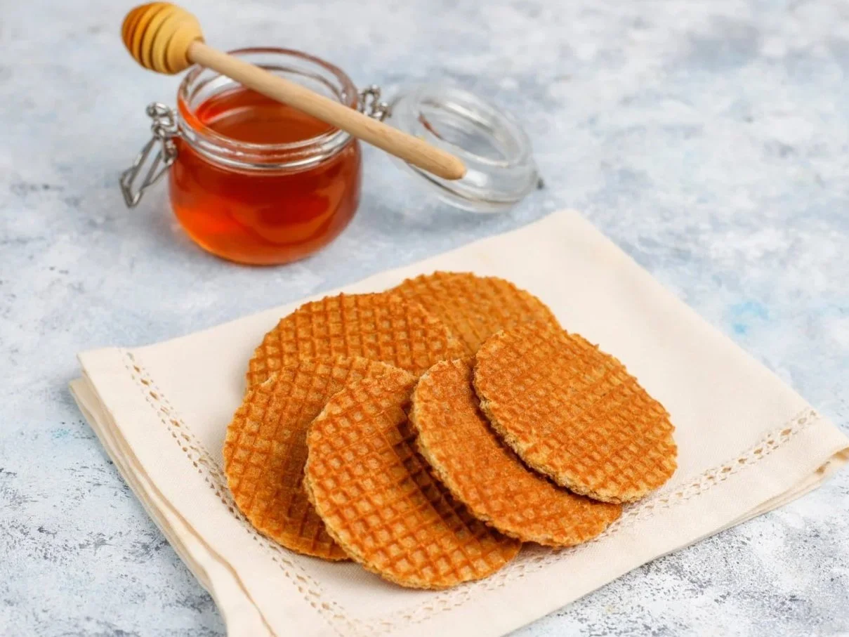 A stack of golden-brown stroopwafels resting on a beige cloth napkin, with a glass jar of honey and a wooden dipper in the background on a textured light-blue surface.