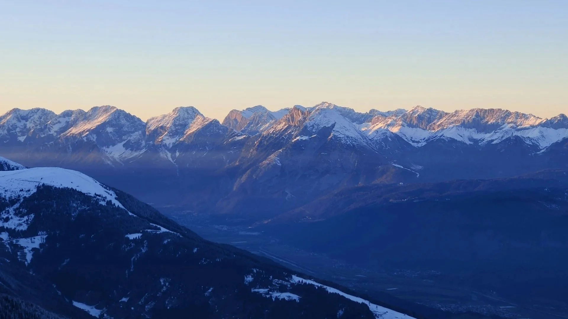 A wide panoramic view of snow-capped mountain peaks under a clear, pale blue sky at dawn or dusk.