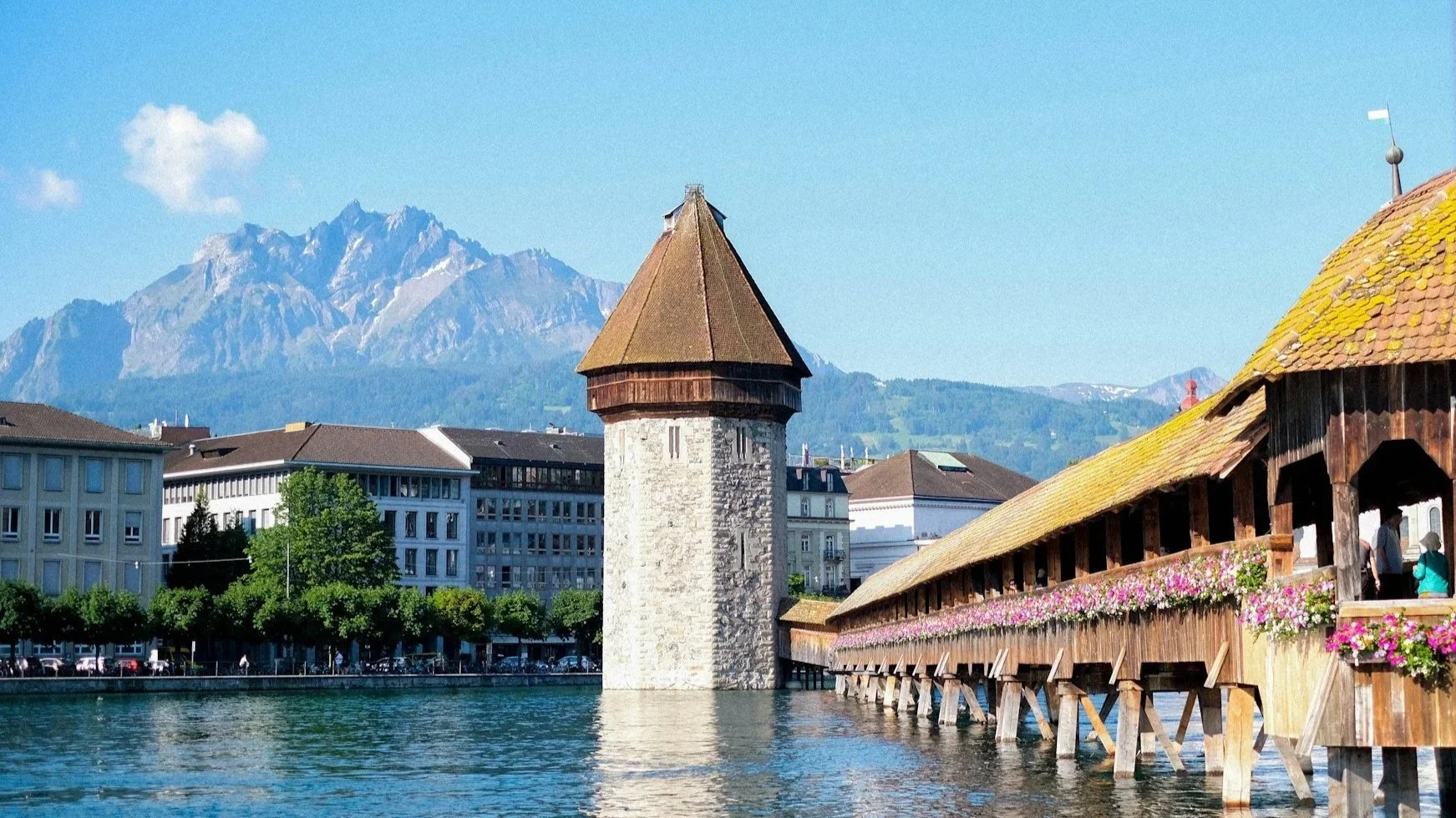 The medieval wooden Chapel Bridge and Water Tower in Lucerne, decorated with pink flowers against a backdrop of blue water and mountains.