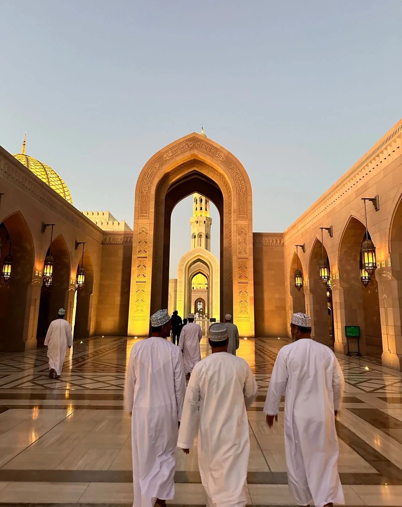 Men in traditional white Omani thobes and embroidered kumma caps walking through the grand arched courtyards of the Sultan Qaboos Grand Mosque at twilight.