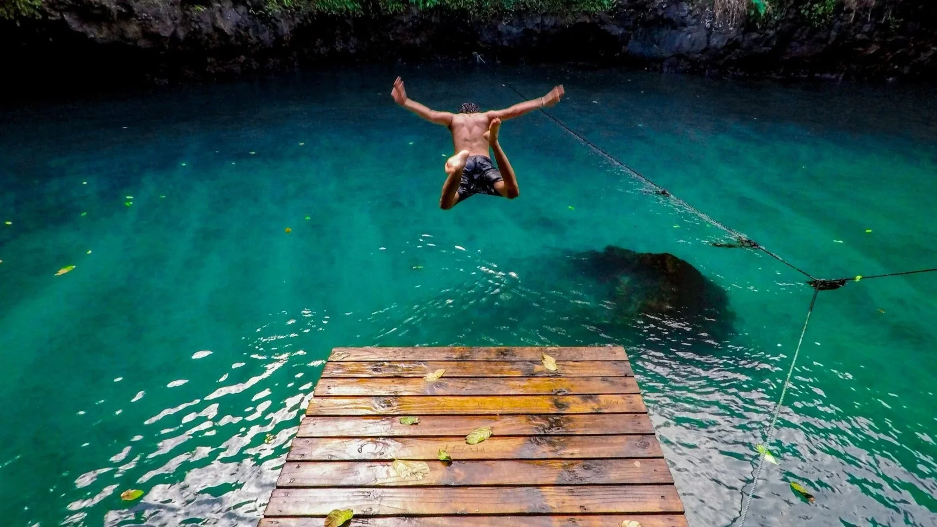 Person leaping off a wooden platform into clear turquoise water surrounded by rocky cliffs.