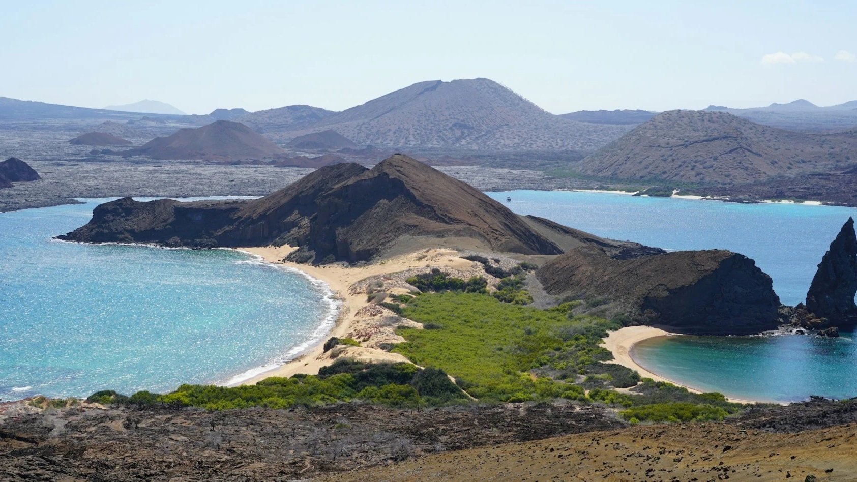 An aerial view of a volcanic island coastline featuring turquoise bays, sandy beaches, green shrubbery, and a tall, sharp rock pinnacle.