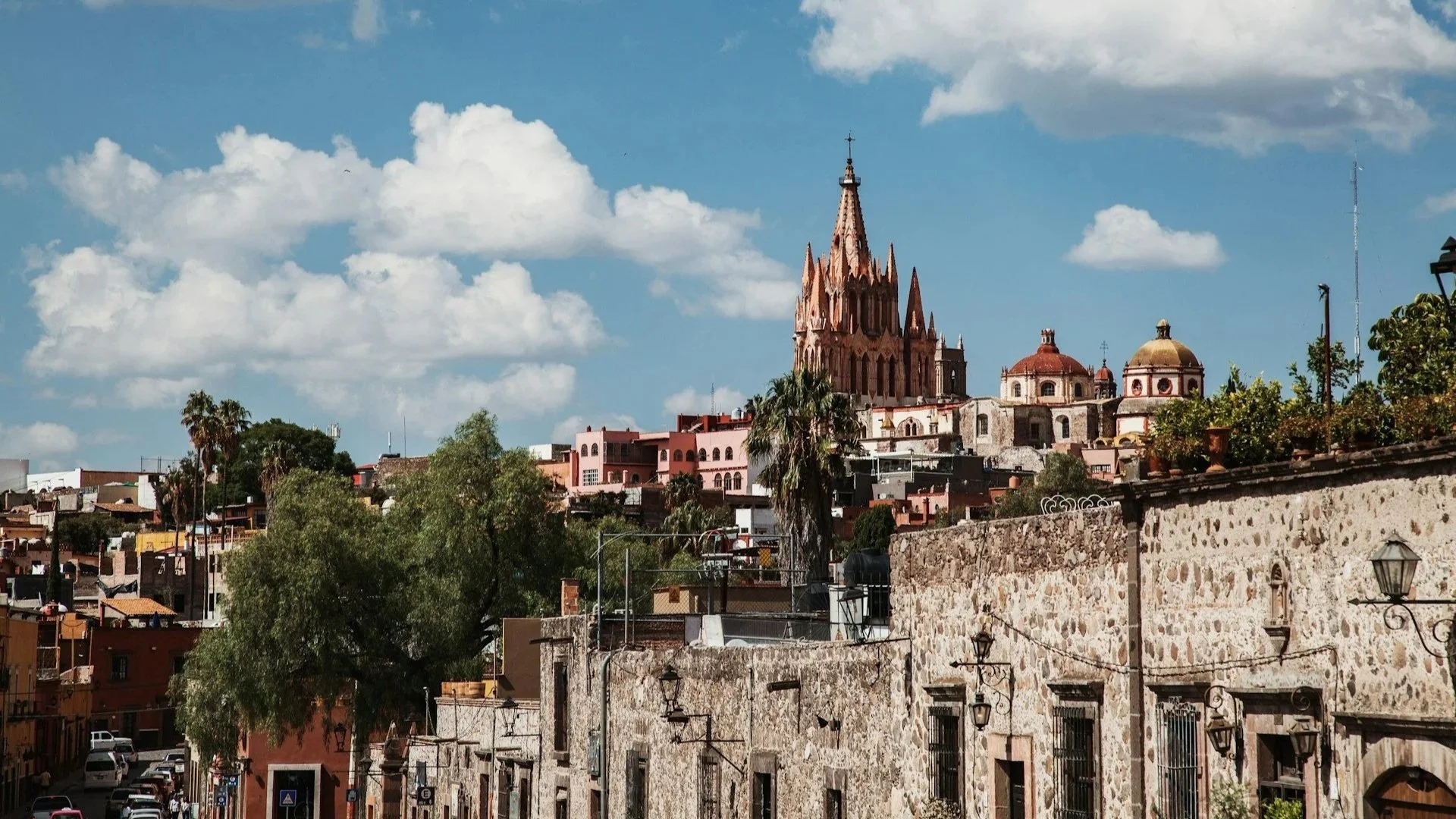 A view of San Miguel de Allende featuring stone buildings in the foreground and the iconic pink, neo-Gothic spires of the Parroquia de San Miguel Arcángel under a blue, cloudy sky.