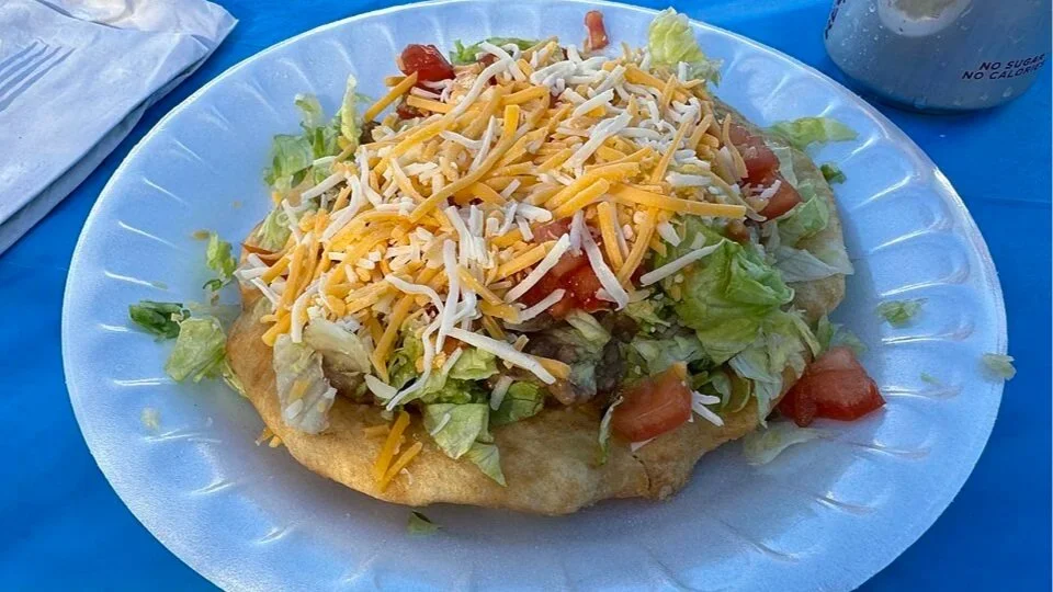 A piece of golden-brown frybread topped with shredded lettuce, diced tomatoes, and cheese served on a white plate.