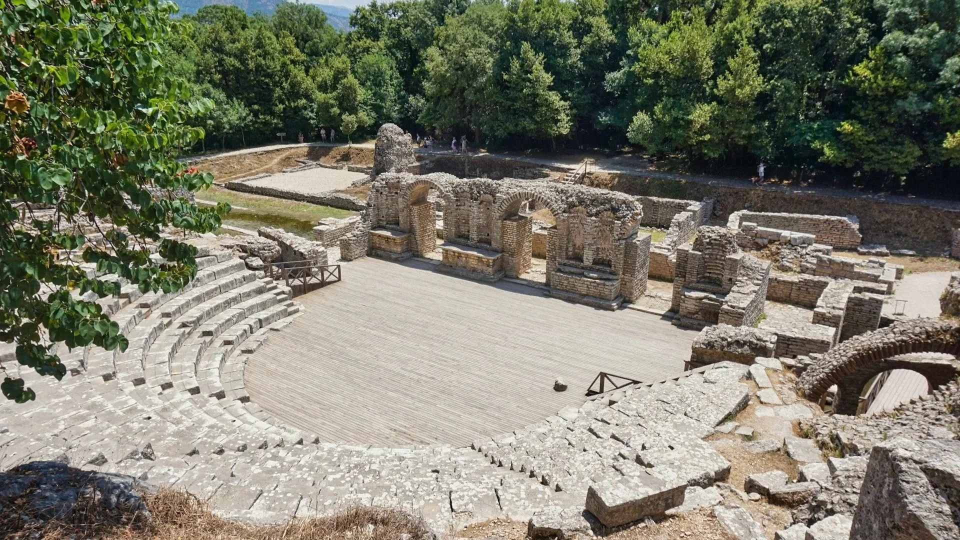 Stone amphitheater ruins surrounded by lush green trees under a bright blue sky.