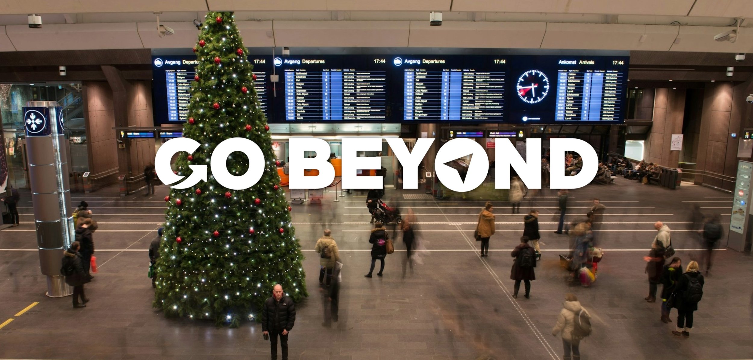 A large, decorated Christmas tree stands in a busy airport terminal under a flight departure board, with the text "GO BEYOND" overlaid in white.