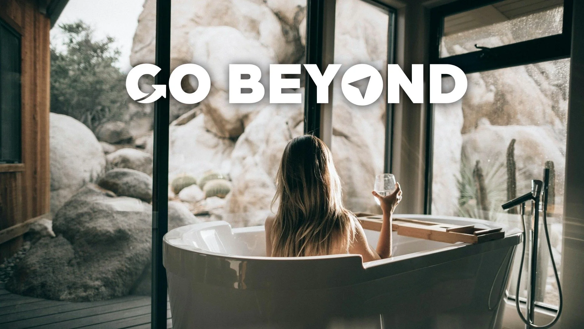 A woman seen from behind relaxes in a white bathtub overlooking a desert landscape, with the text "GO BEYOND" overlaid in large white letters across the center.