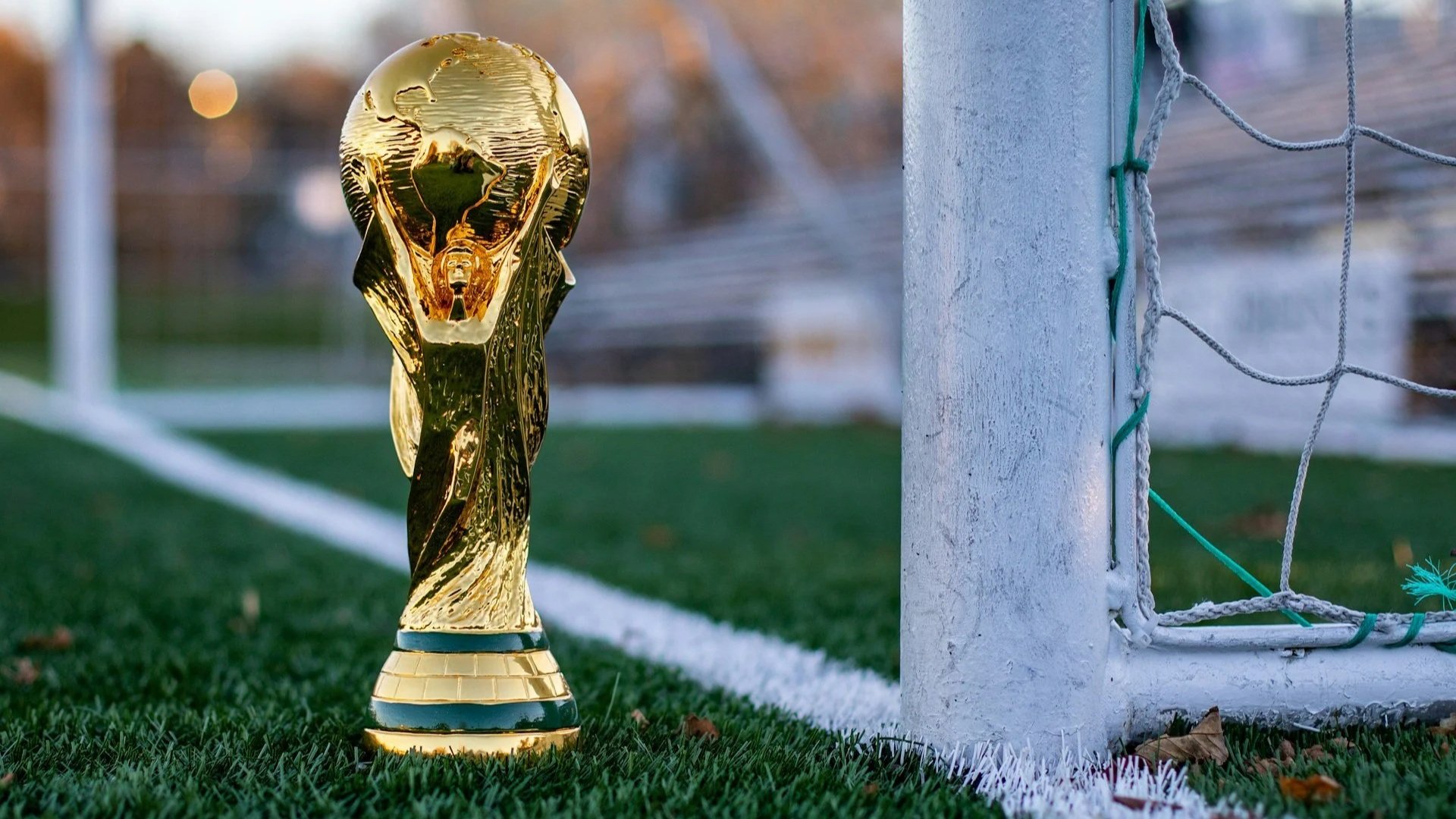 A close-up of a gold World Cup-style trophy standing on the green grass of a pitch next to a white goalpost.