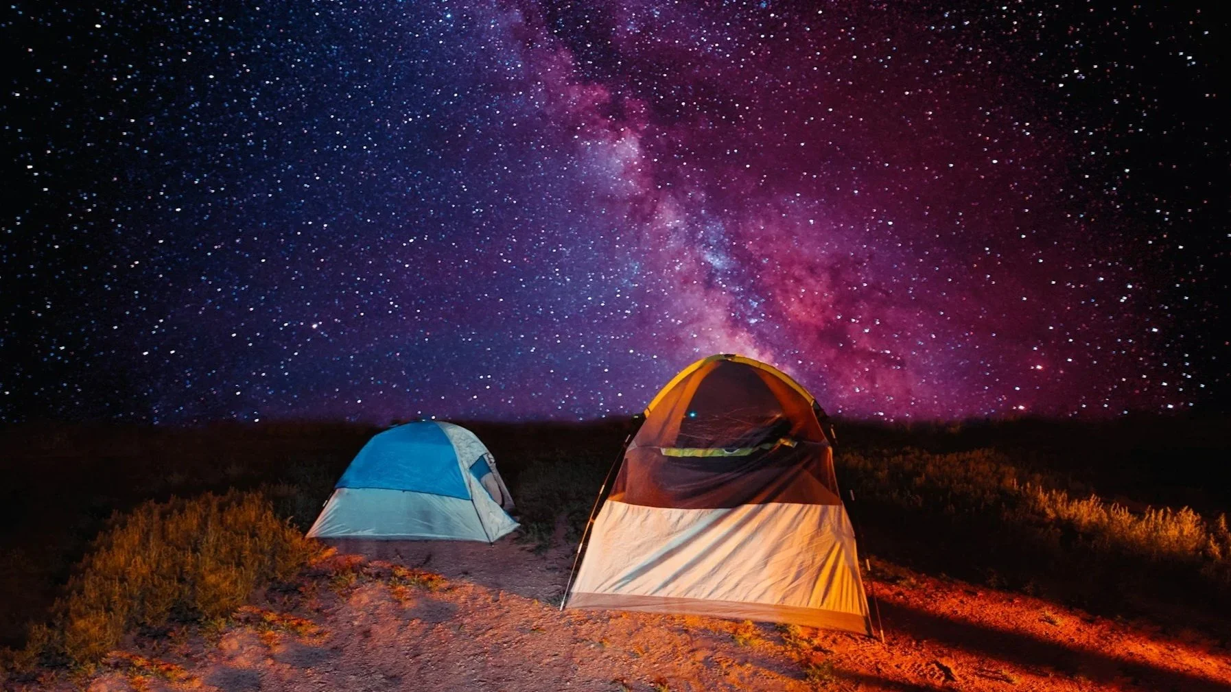A photograph of two illuminated tents at a campsite under a vibrant, purple-hued Milky Way galaxy and a dense field of stars.