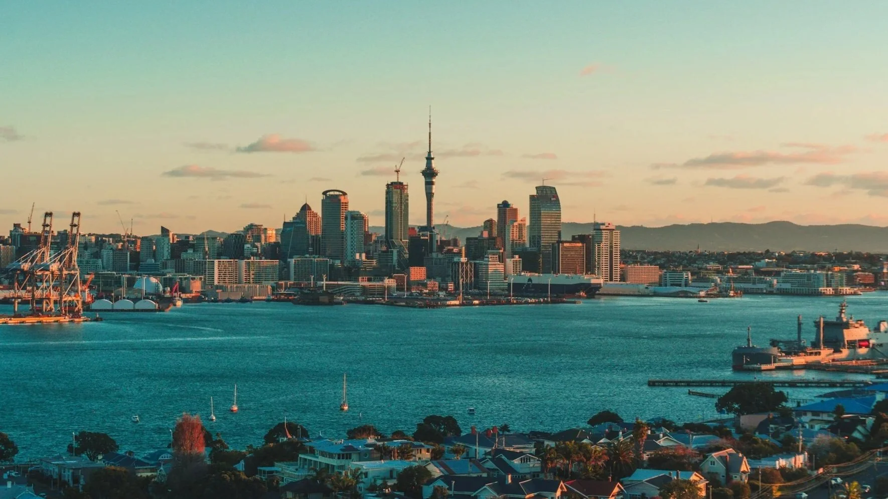 The Auckland city skyline and Sky Tower at sunset, viewed across the harbor from a distance.