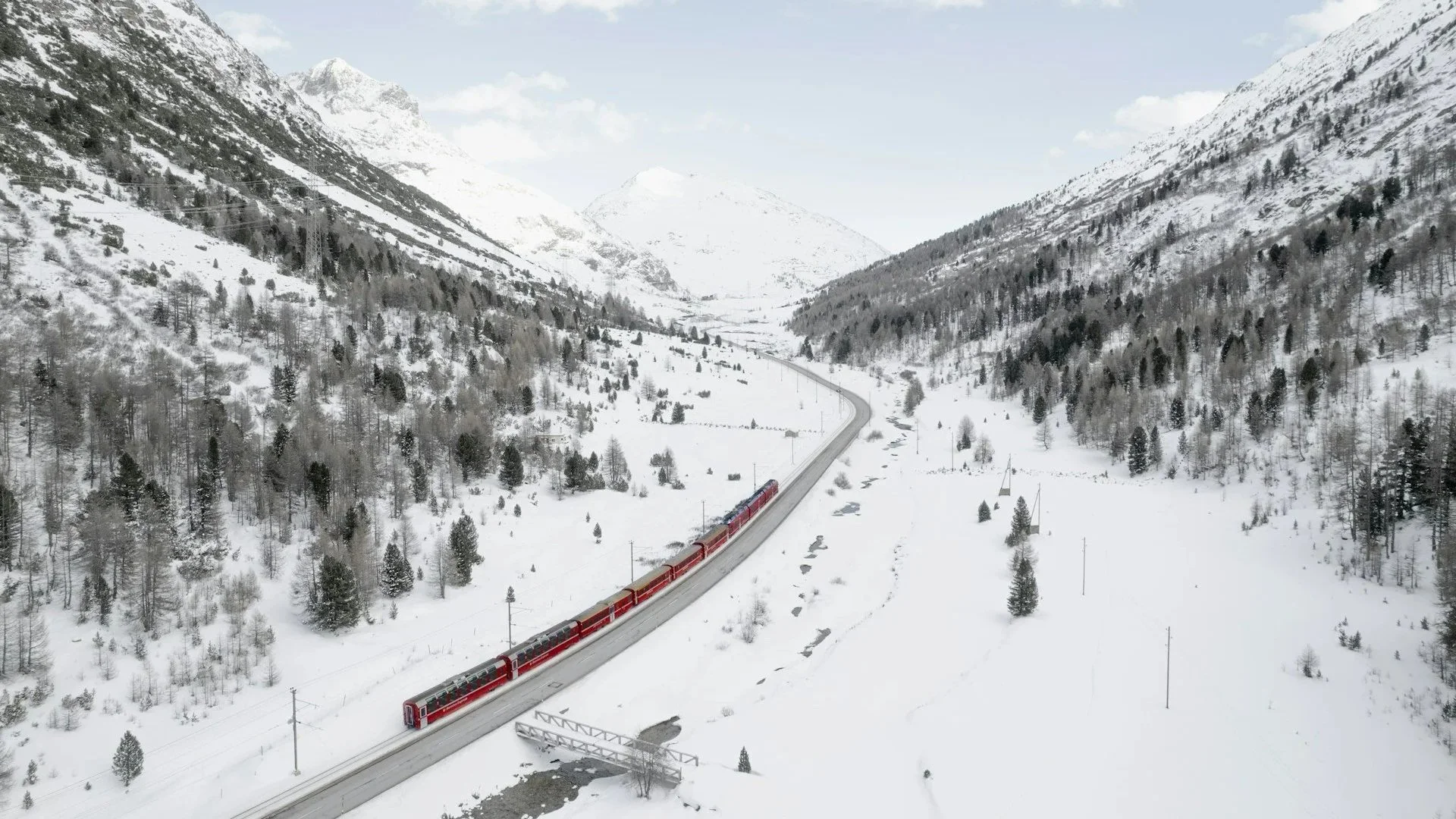 A long red train traveling through a snow-covered valley surrounded by white, pine-covered mountains under a bright sky.