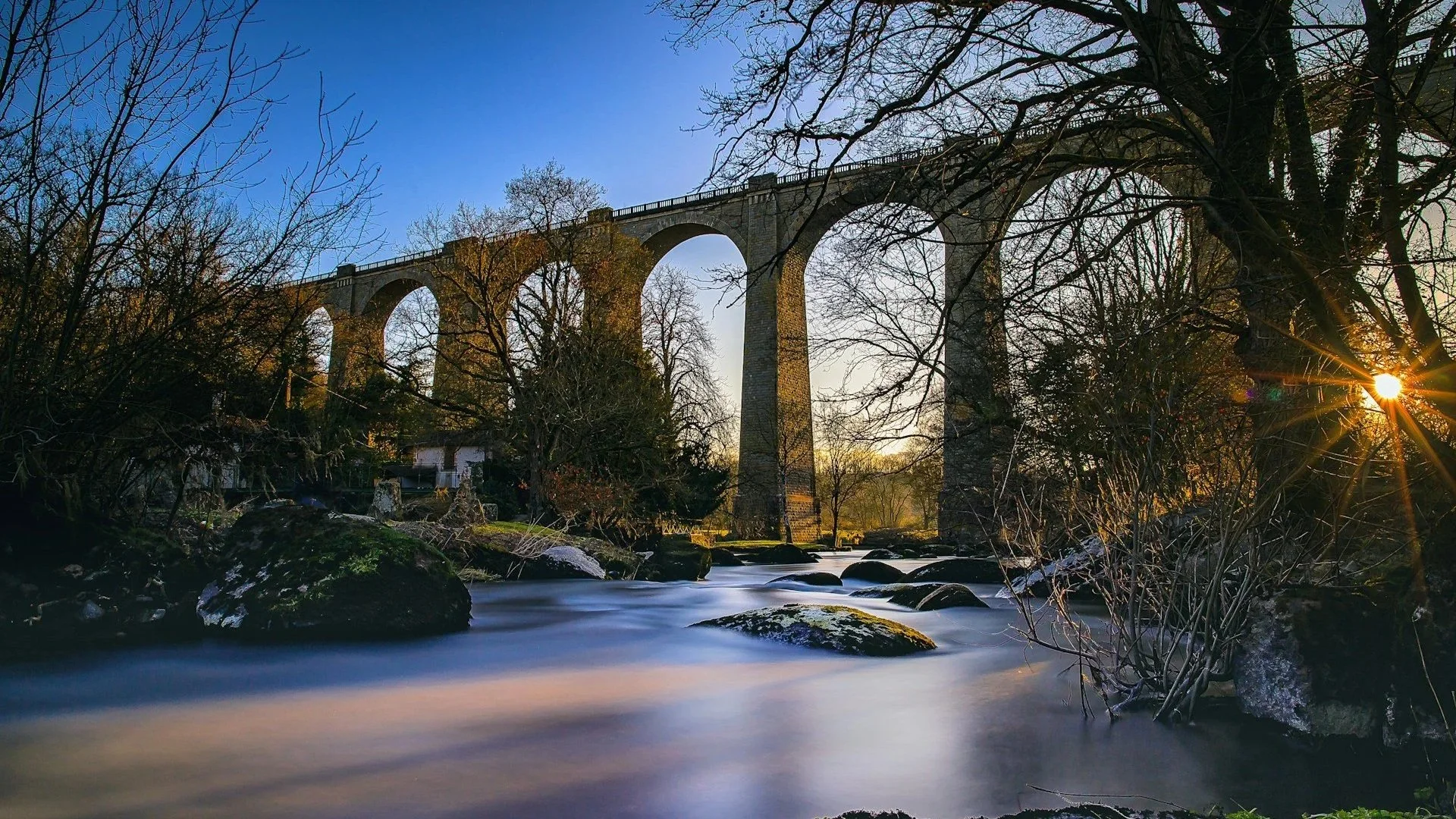 A tall stone arch viaduct bridge spanning across a river at sunset, with bare winter trees in the foreground.