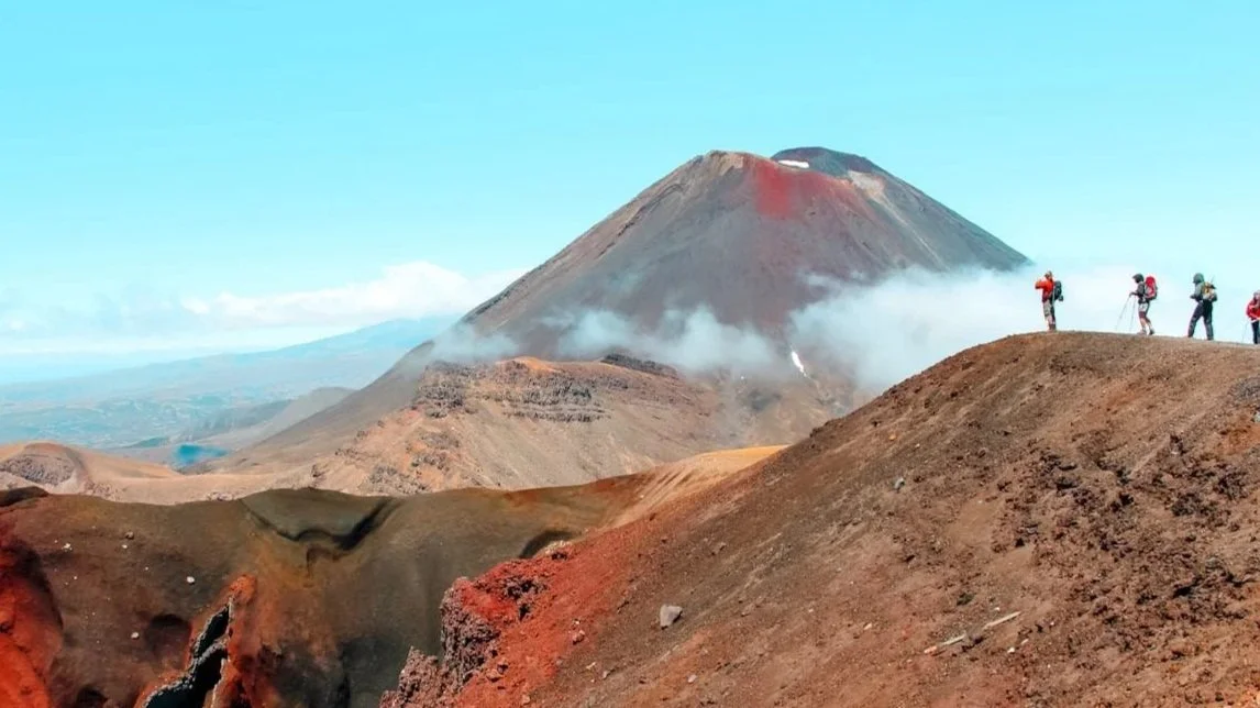 A group of hikers stands on a red volcanic ridge overlooking the massive cone of Mount Ngauruhoe in Tongariro National Park.