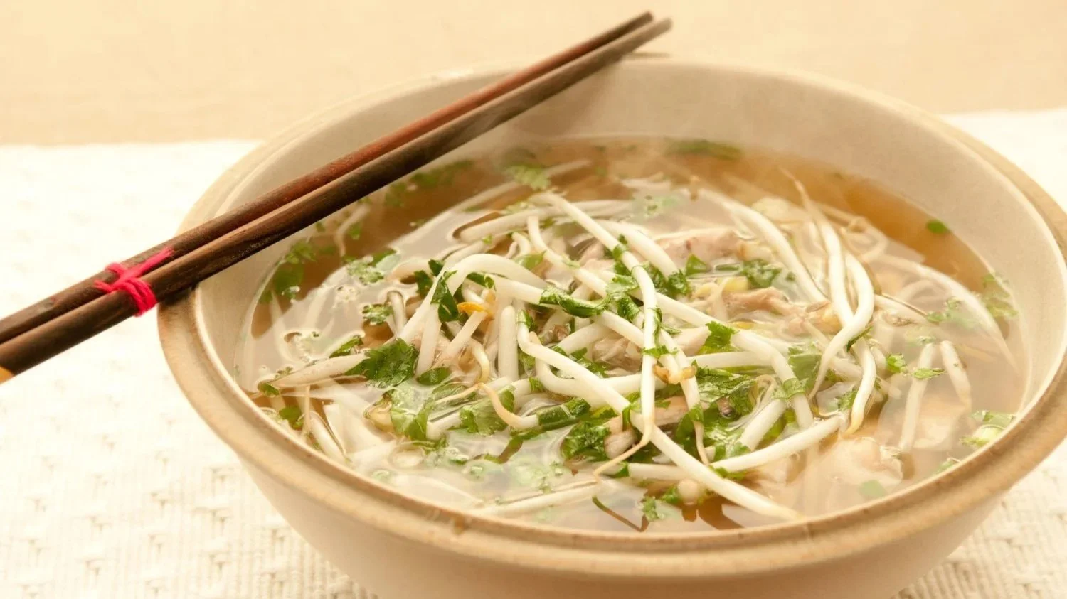 A steaming bowl of Vietnamese Pho with clear broth, rice noodles, fresh bean sprouts, and herbs, served with wooden chopsticks.
