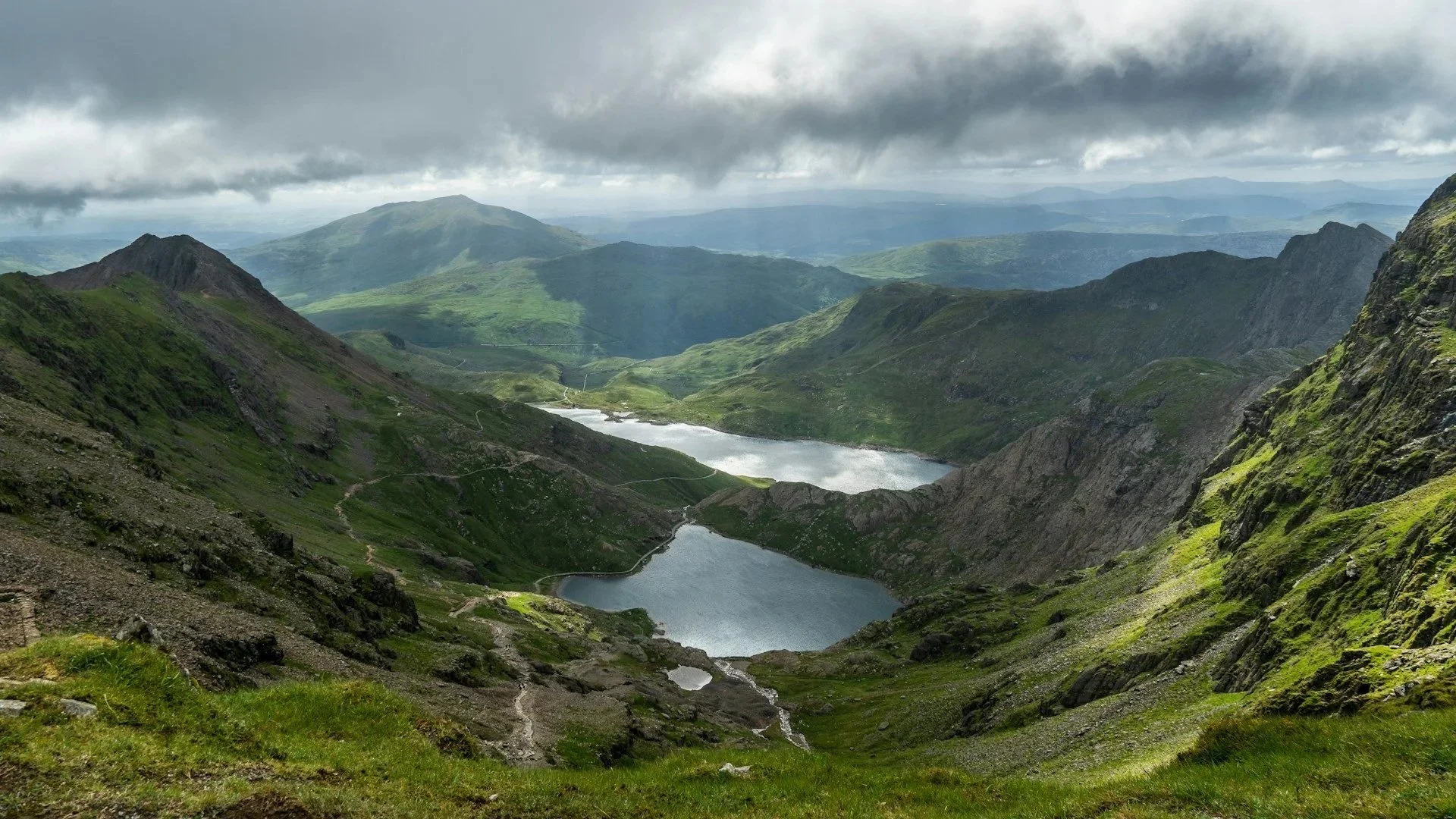 A wide valley view between green, rocky mountains under a cloudy sky, showcasing two bright blue lakes below.