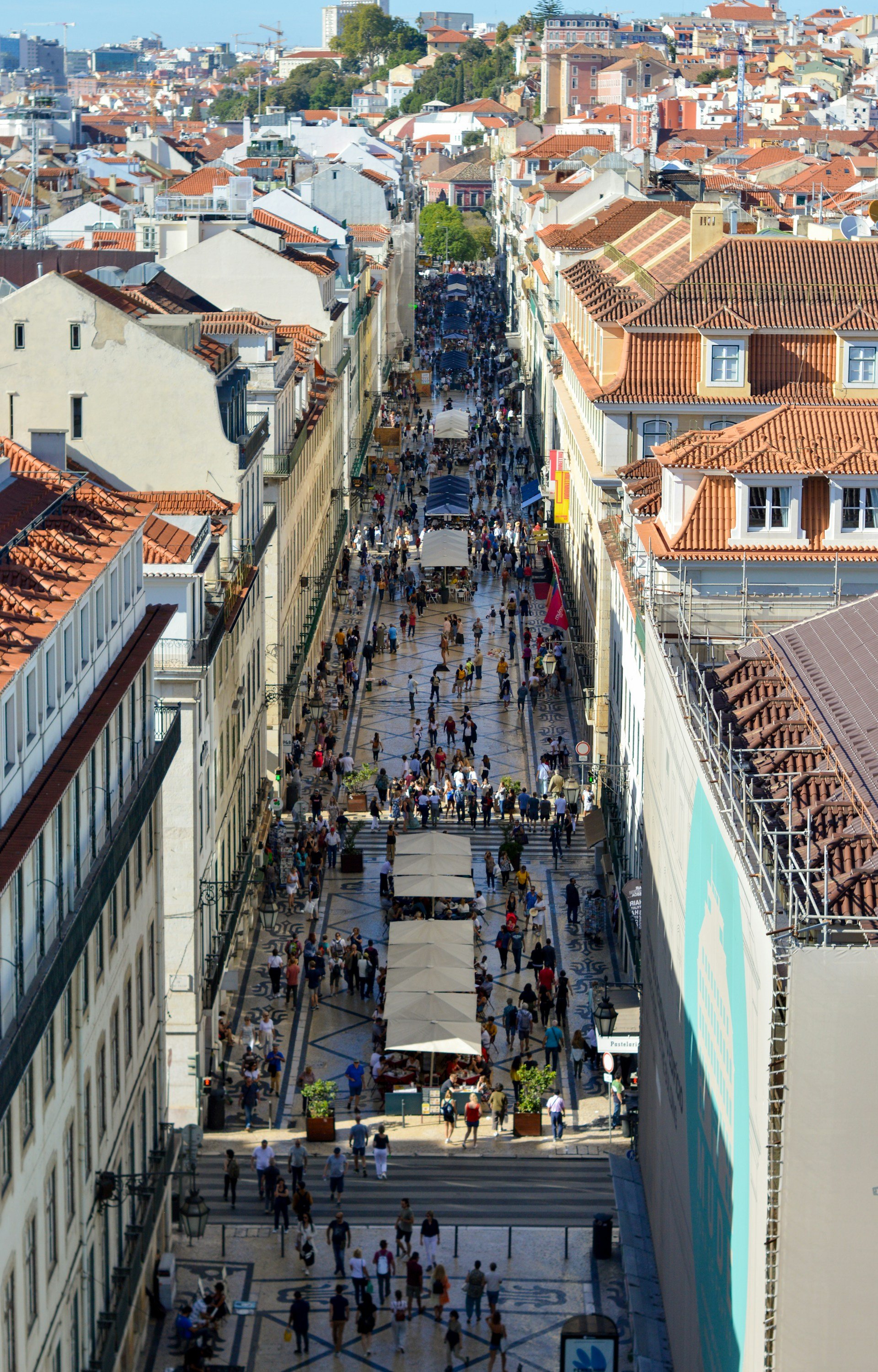 A high-angle view of a crowded pedestrian street in Lisbon, lined with traditional buildings and red-tiled roofs.