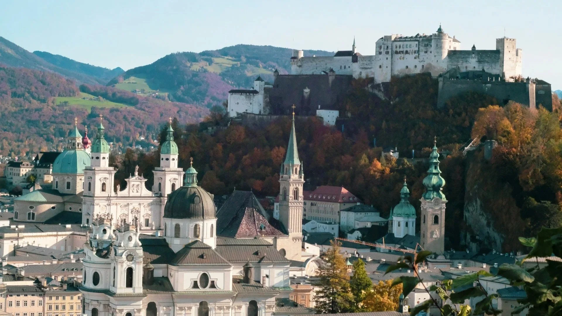 A high-angle view of Salzburg's skyline, showing several green-domed churches and the Hohensalzburg Fortress sitting atop a hill.