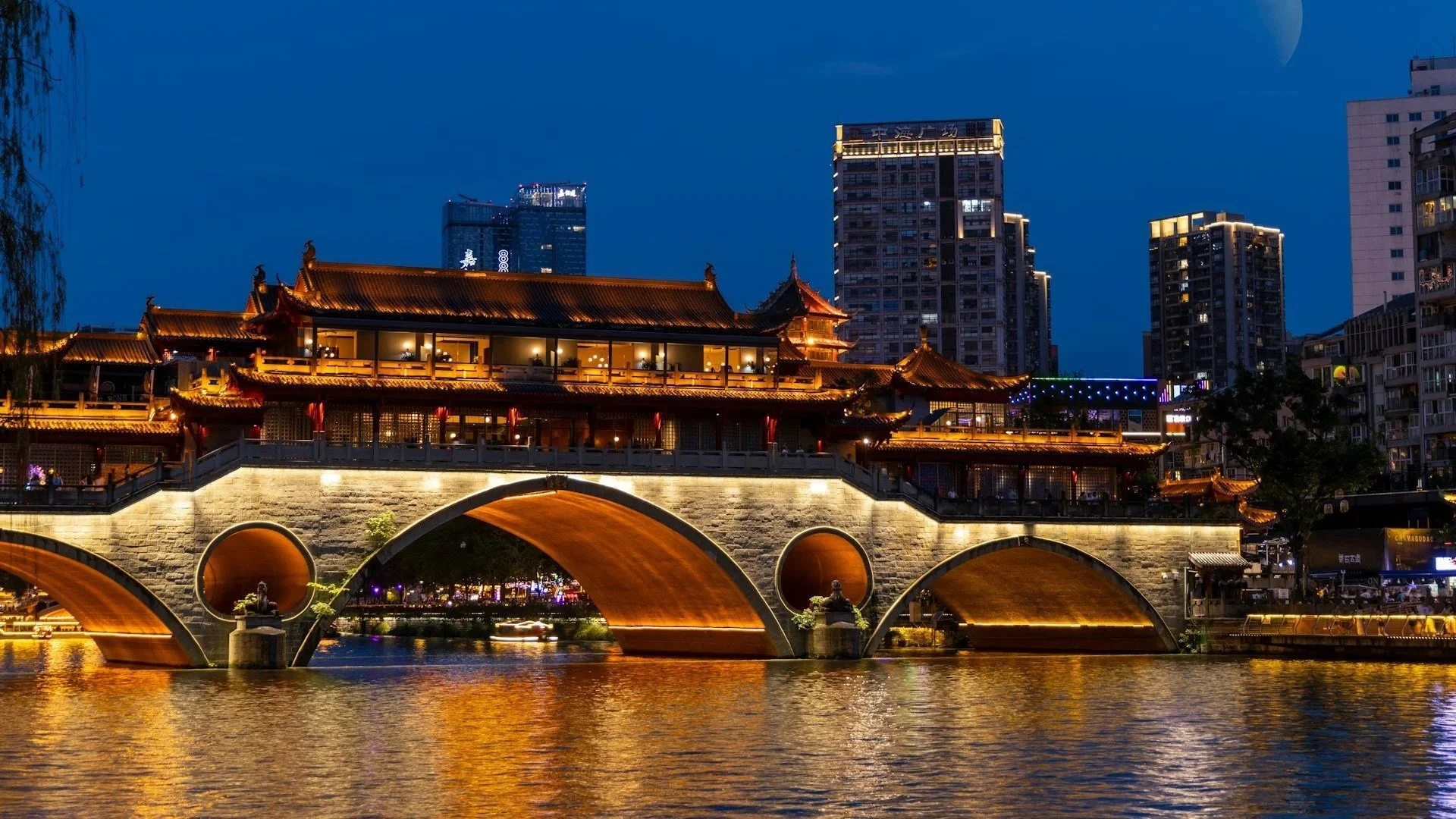Illuminated Anshun Bridge over a river at night with a crescent moon and modern city skyscrapers in the background.