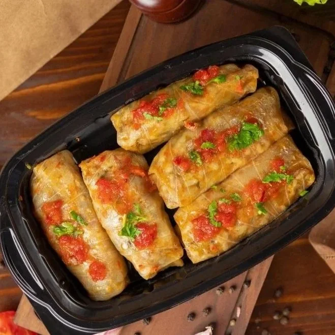 A top-down view of cooked cabbage rolls topped with tomato sauce and fresh herbs inside a black container, set on a rustic wooden table.