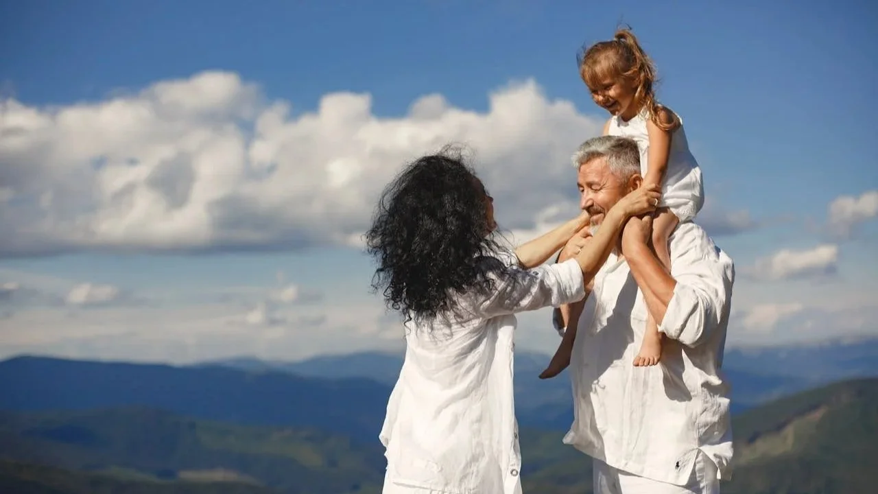A happy family of three dressed in white standing on a grassy hill with a scenic mountain range and blue sky in the background.