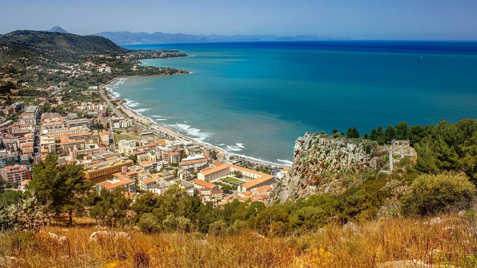High-angle panoramic view of a Mediterranean coastal town with red-roofed buildings nestled between a rocky cliff and a bright blue sea.