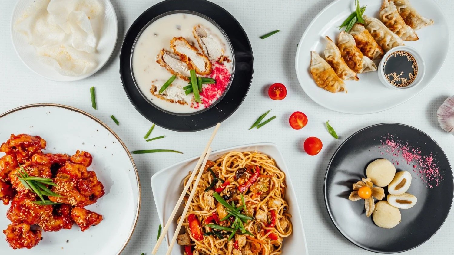 Flat-lay arrangement of various Asian dishes including stir-fried noodles, crispy chicken, dumplings, and dessert.