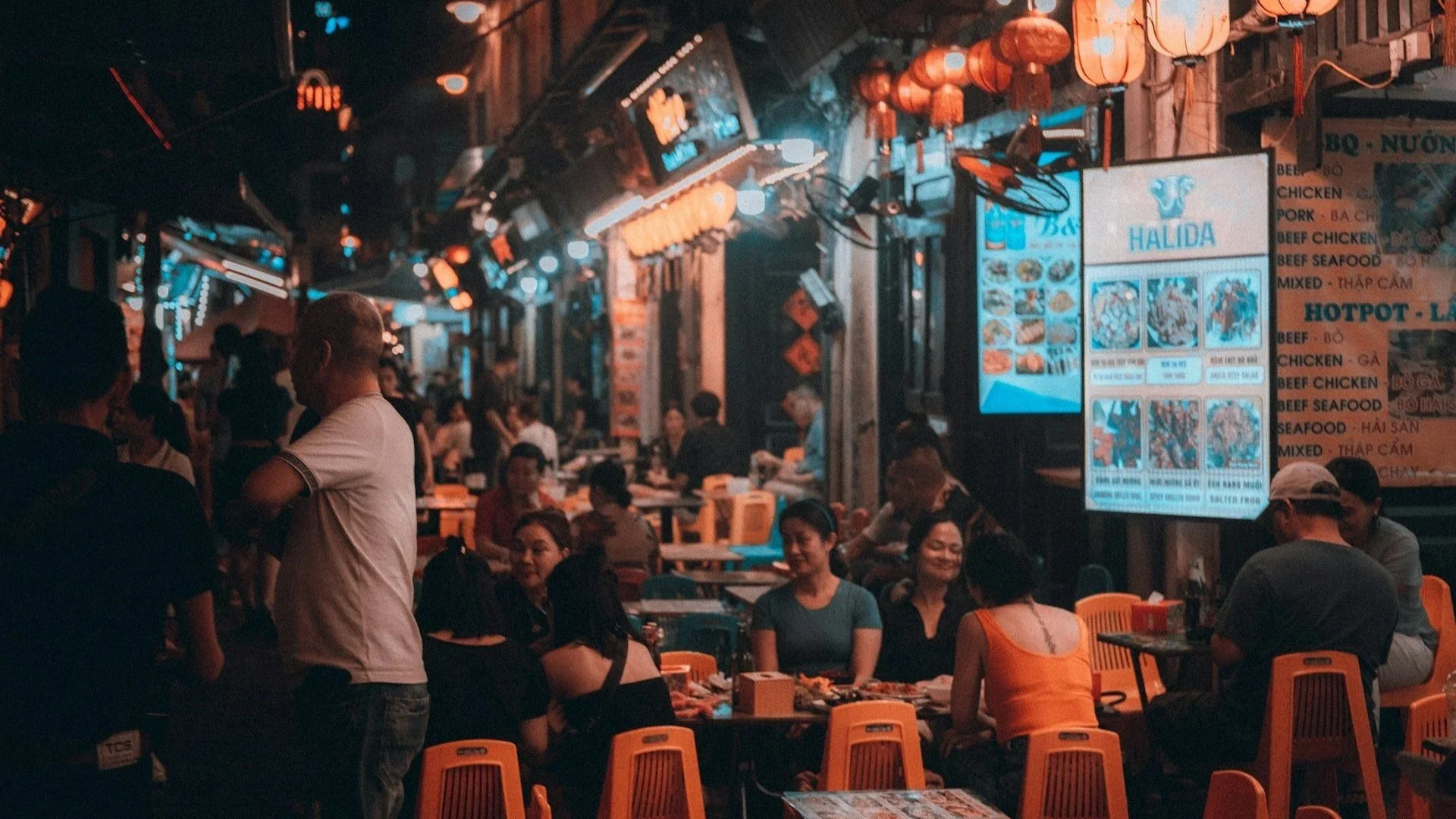 A crowded outdoor street food market in Vietnam at night, lit by warm orange lanterns and neon signs.