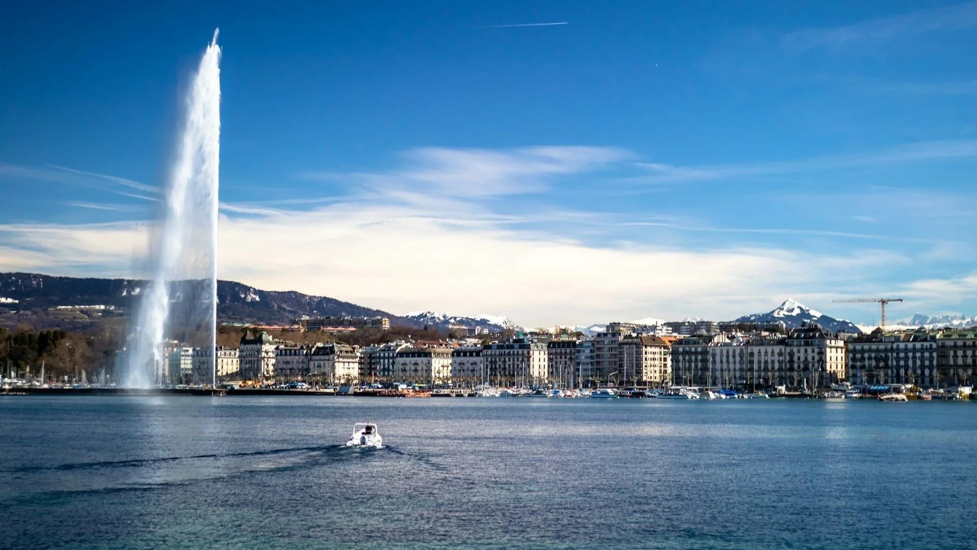 A wide shot of the Jet d'Eau fountain shooting water high into the air on Lake Geneva, with city buildings and snow-capped mountains in the background.