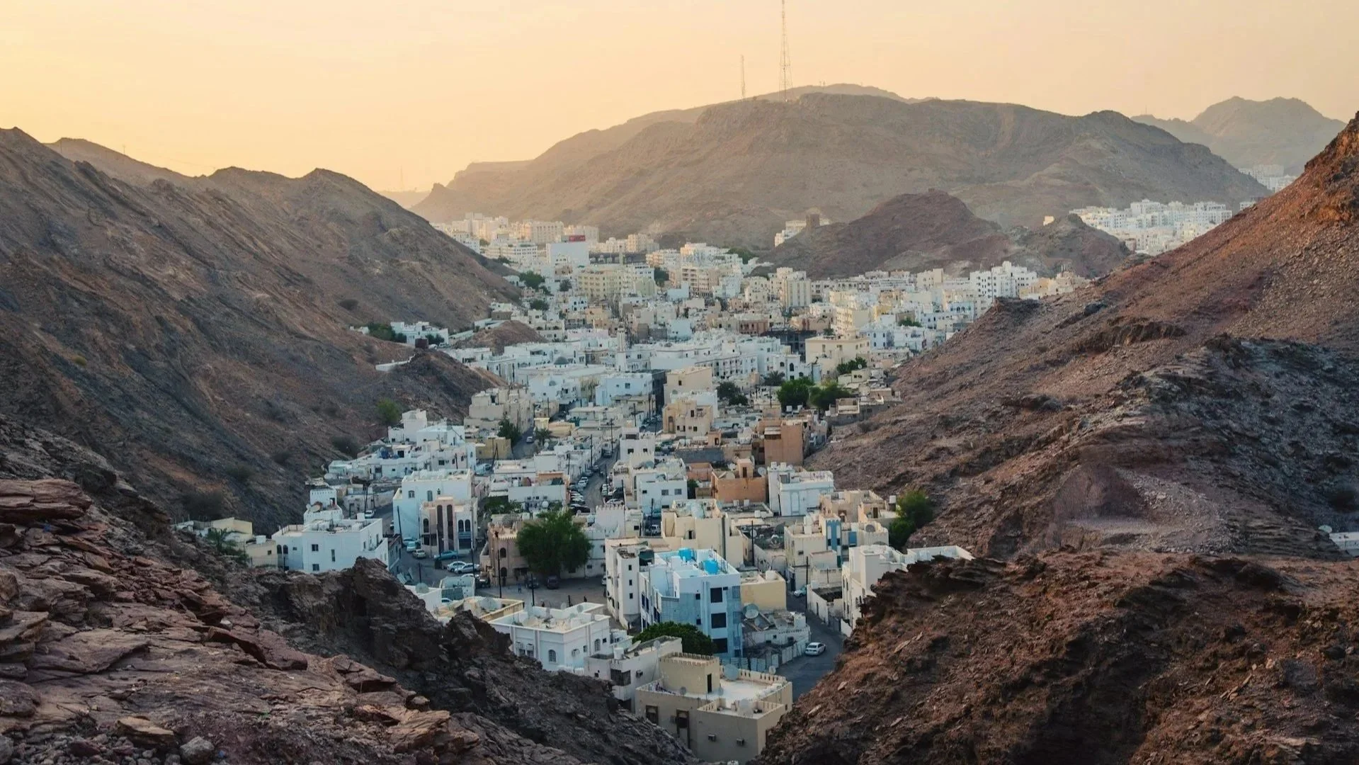 A white-walled town nestled in a narrow valley between towering, rugged mountains at sunset.