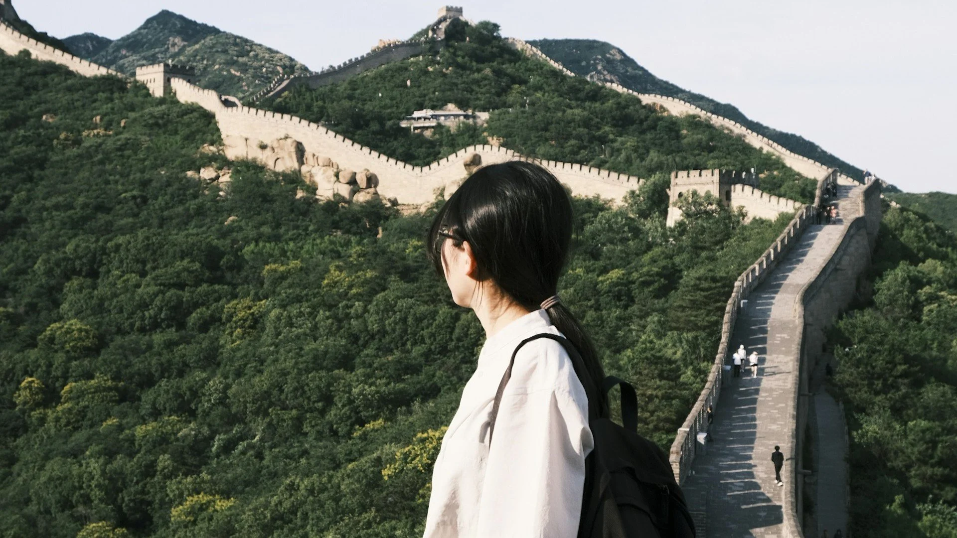 Side profile of a woman with a backpack looking out at the Great Wall of China winding across lush green mountains.