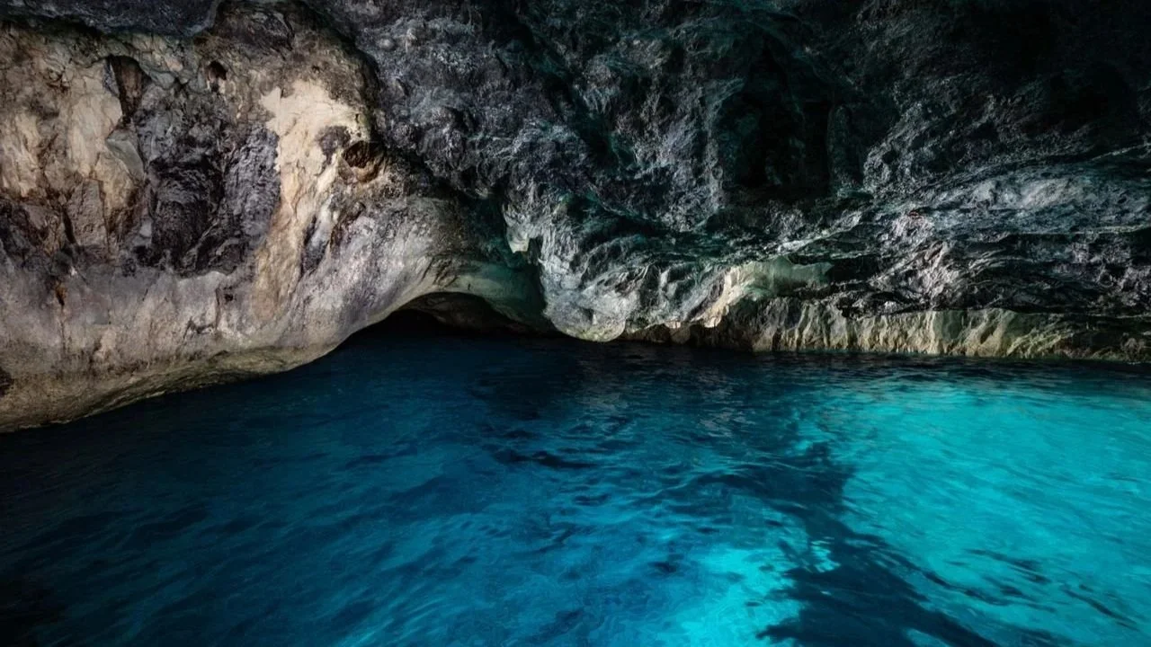 The interior of a dark, rocky sea cave with vibrant, glowing turquoise water reflecting light onto the stone ceiling.