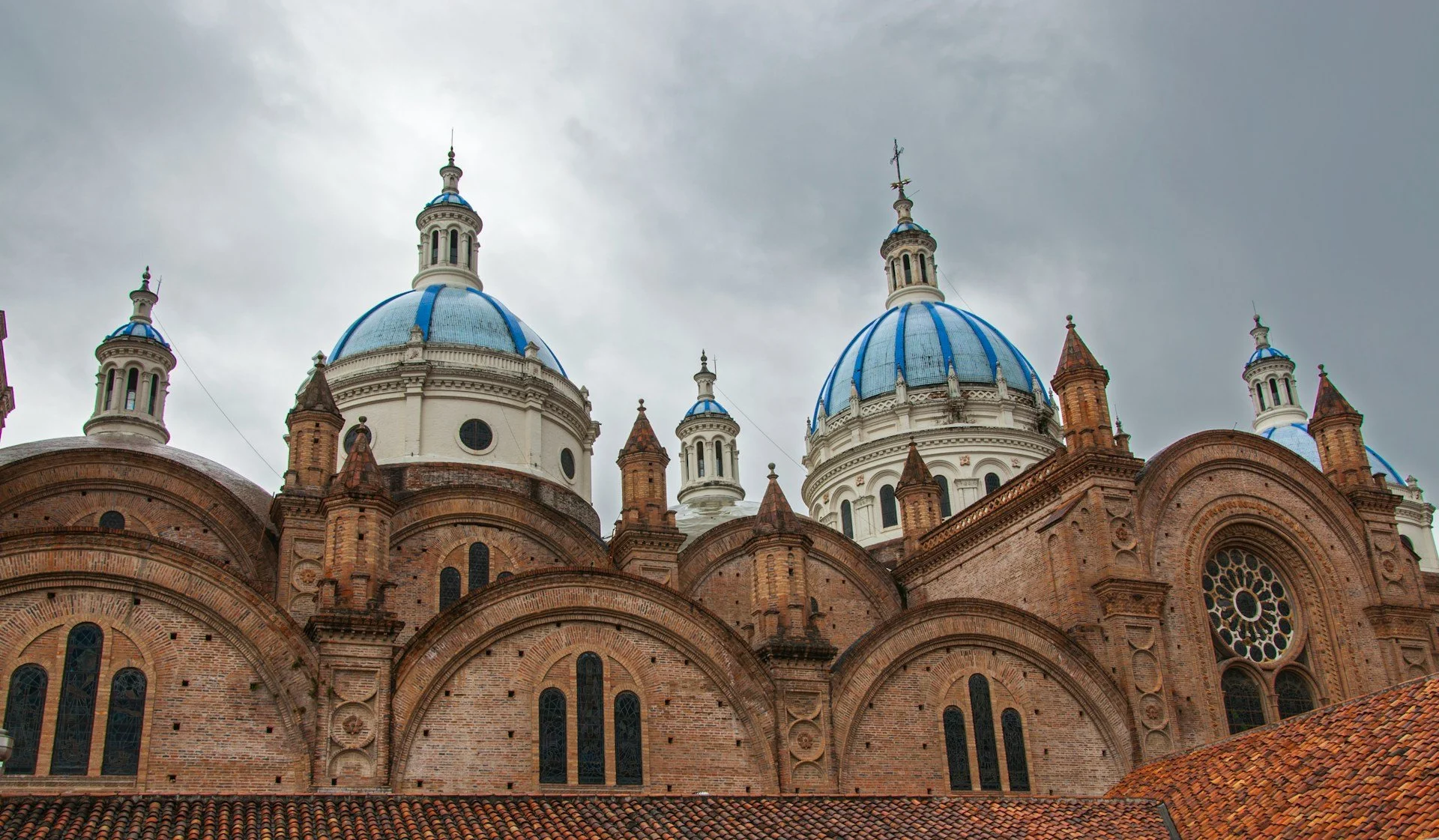 The upper exterior of the New Cathedral in Cuenca, showing its intricate red brickwork and signature bright blue and white tiled domes.