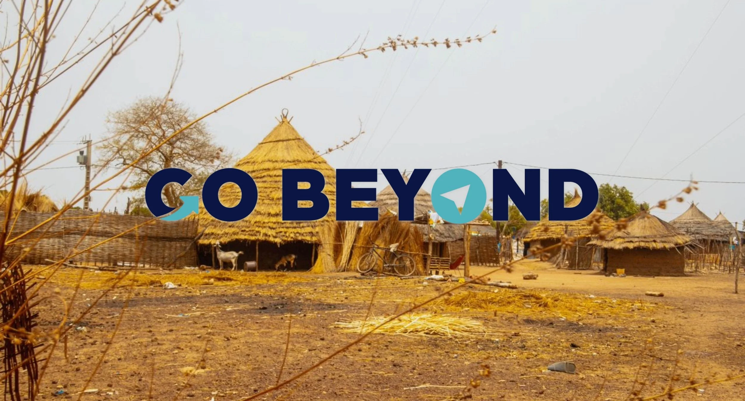 Traditional thatched-roof huts in a rural village setting, overlaid with a large dark blue text logo that reads "GO BEYOND".