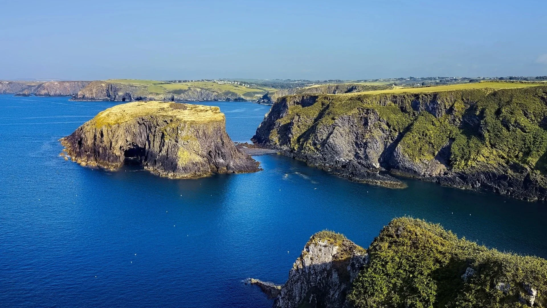 High-angle view of rugged green coastal cliffs meeting a deep blue ocean, with a prominent rocky islet just off the shore.