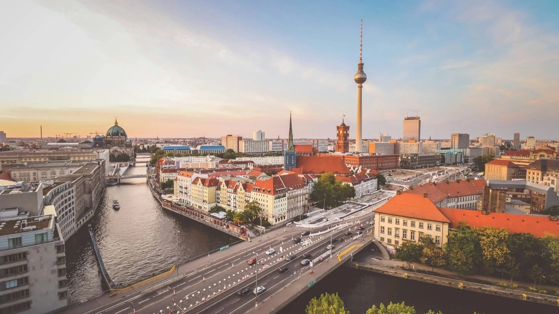 Scenic evening view of Berlin, Germany, showing a multi-lane bridge crossing the Spree River, with the iconic Berlin TV Tower (Fernsehturm) and the dome of the Berlin Cathedral in the background.