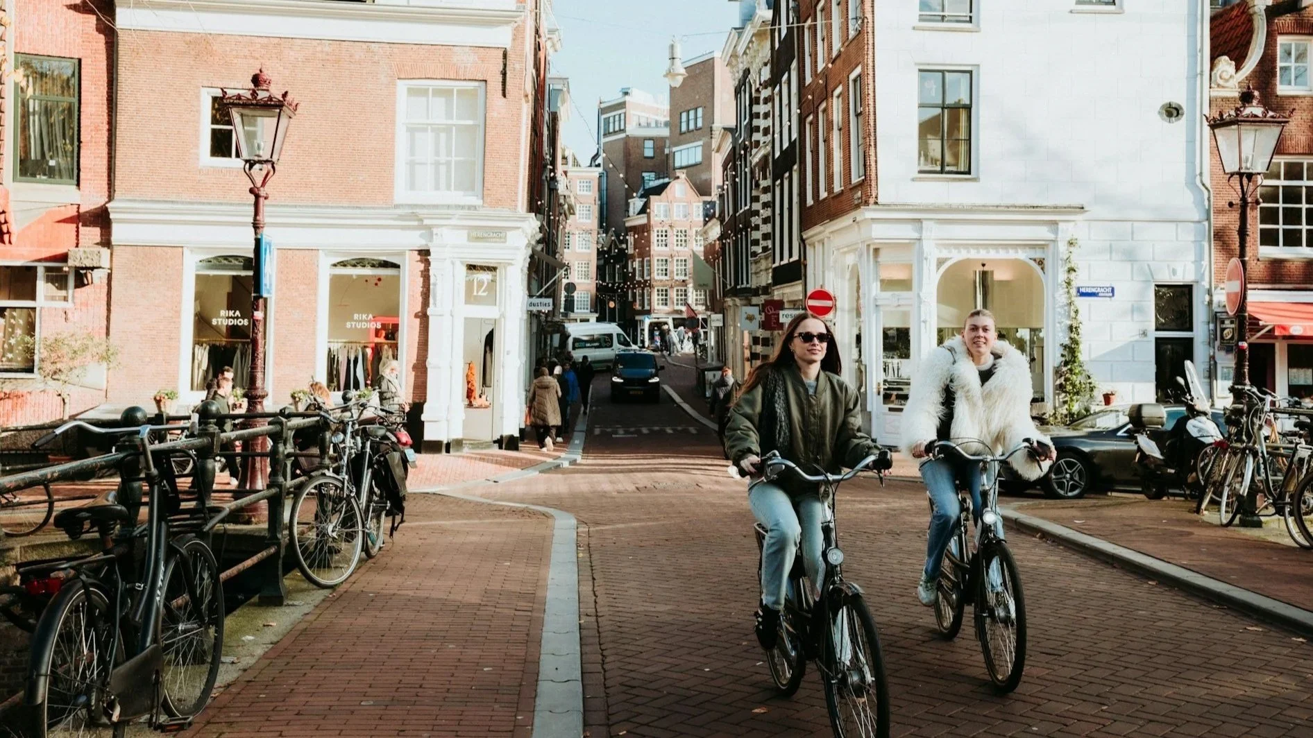 Two people cycling down a sunlit Amsterdam street lined with historic brick buildings and boutiques.