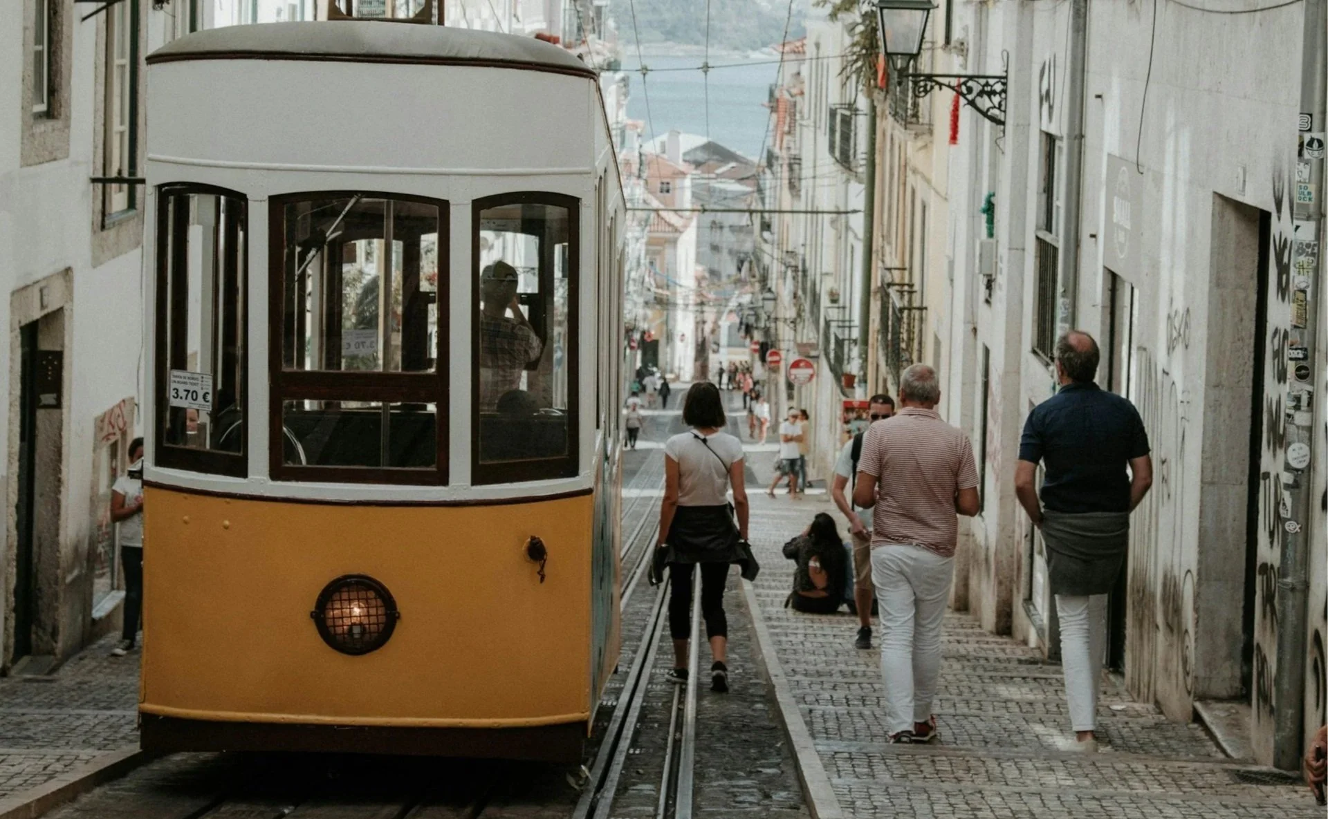 An iconic yellow and white funicular climbing a steep, narrow street in Lisbon, with people walking on the adjacent cobblestone sidewalk.