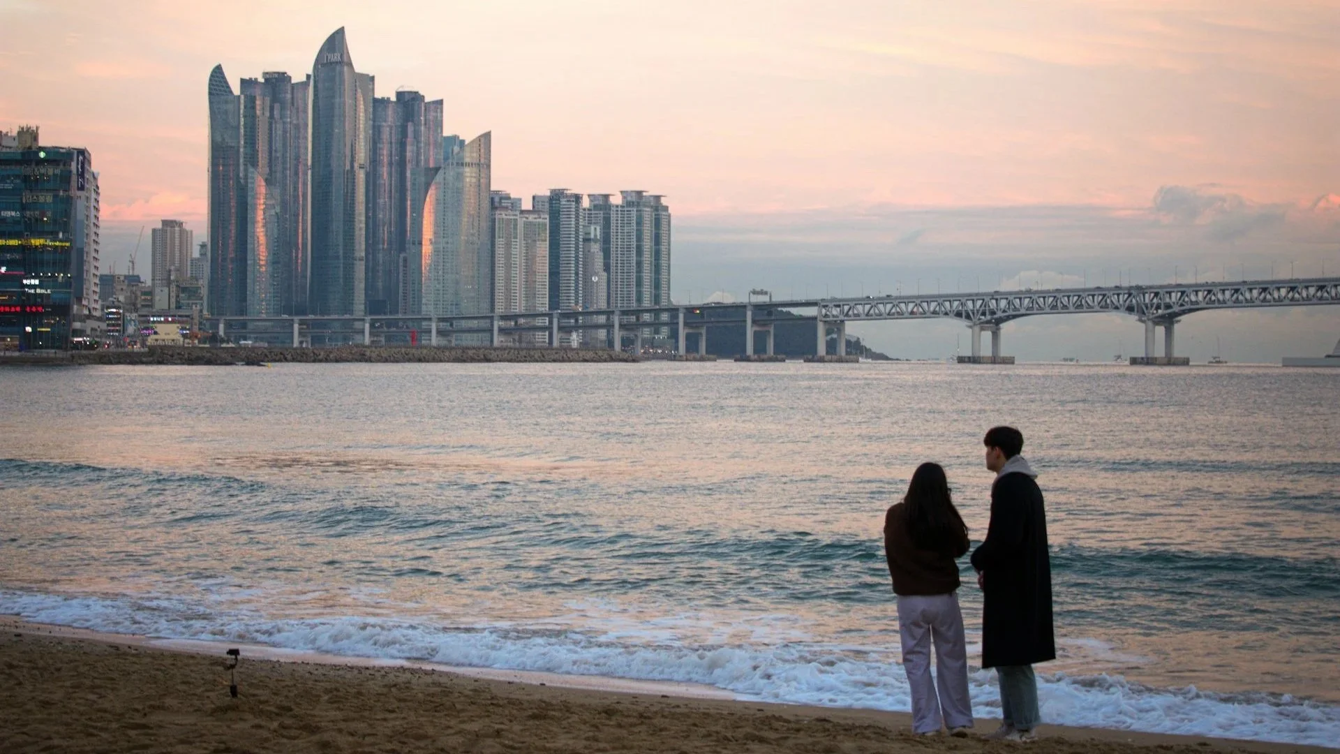 A couple stands on a sandy beach looking toward a cluster of modern glass skyscrapers and a long white suspension bridge over the water at dusk.